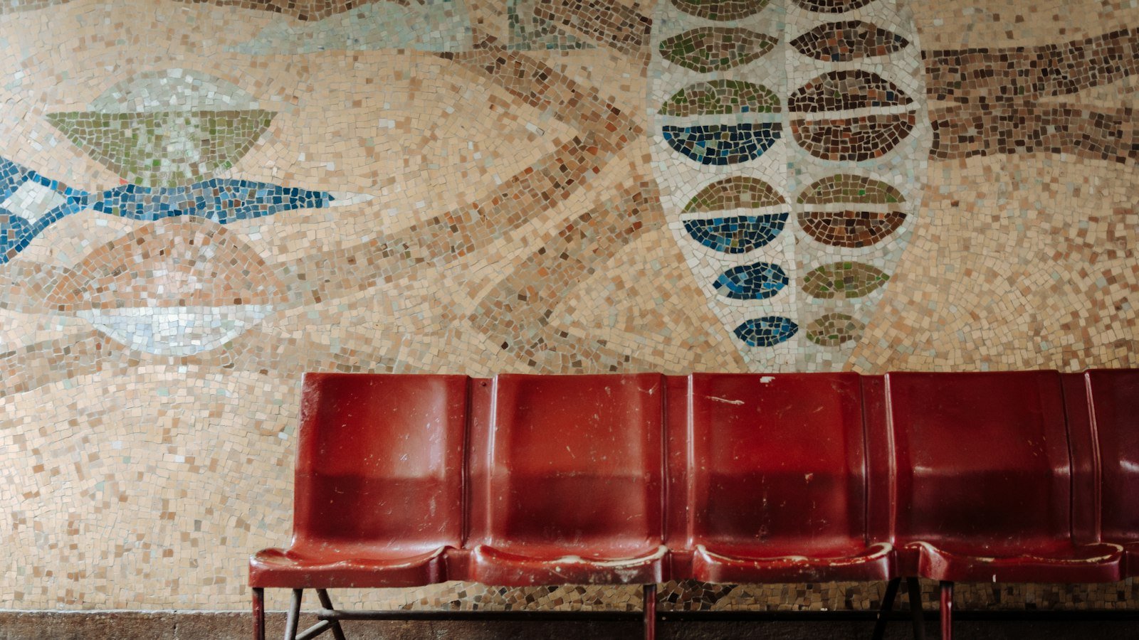A row of empty red waiting-room chairs against a mosaic wall