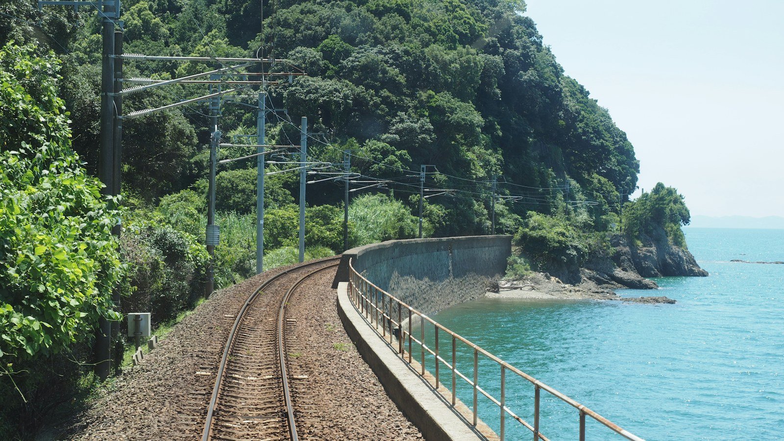 Railway track curving along a green coastline beside a blue sea
