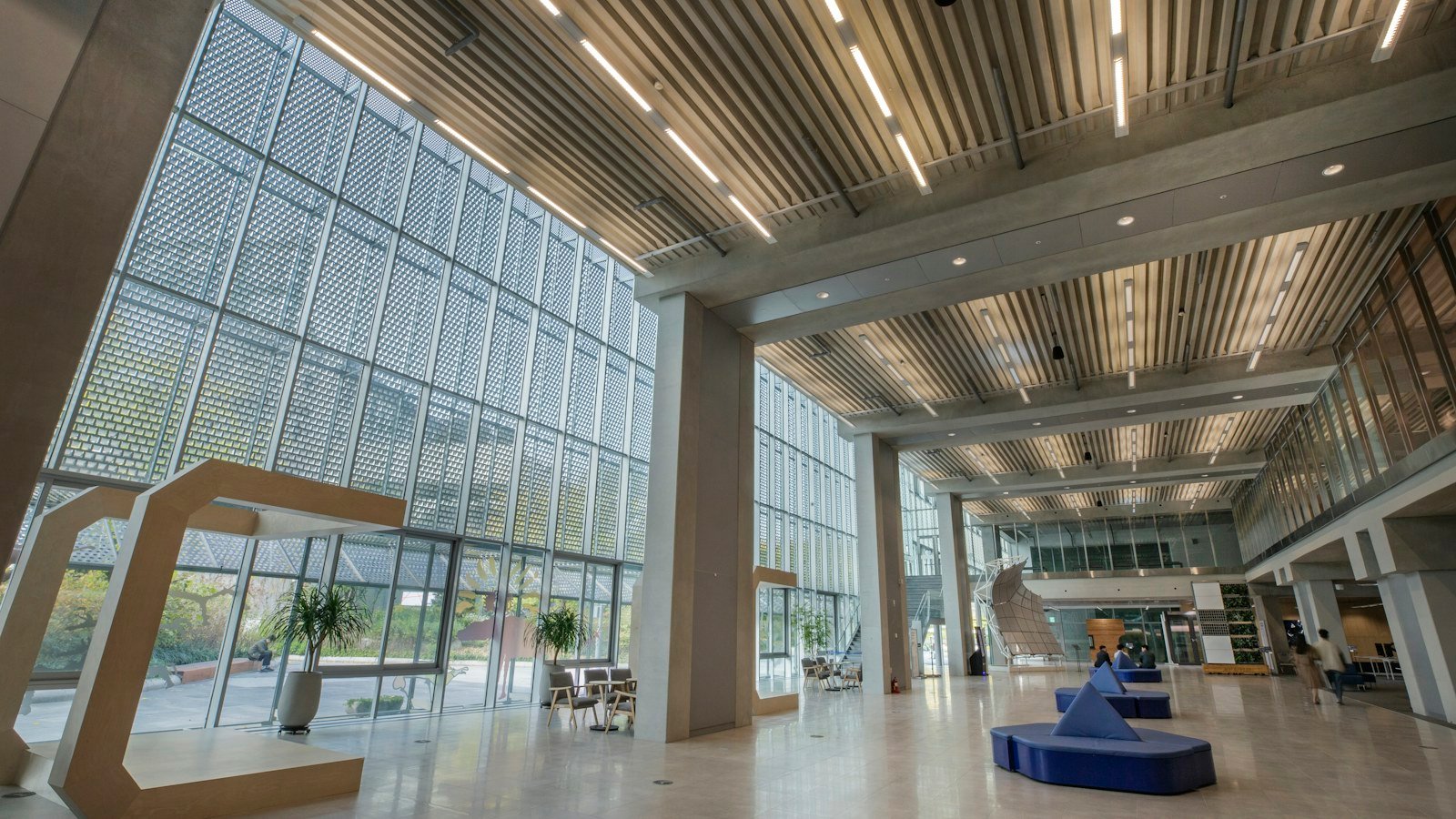 Bright, mostly empty atrium with tall glass walls and blue seating